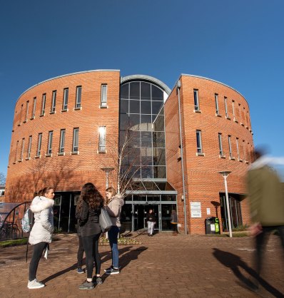 A group of people stand and walk near a modern, curved brick building with large windows at Chester University under a clear blue sky.