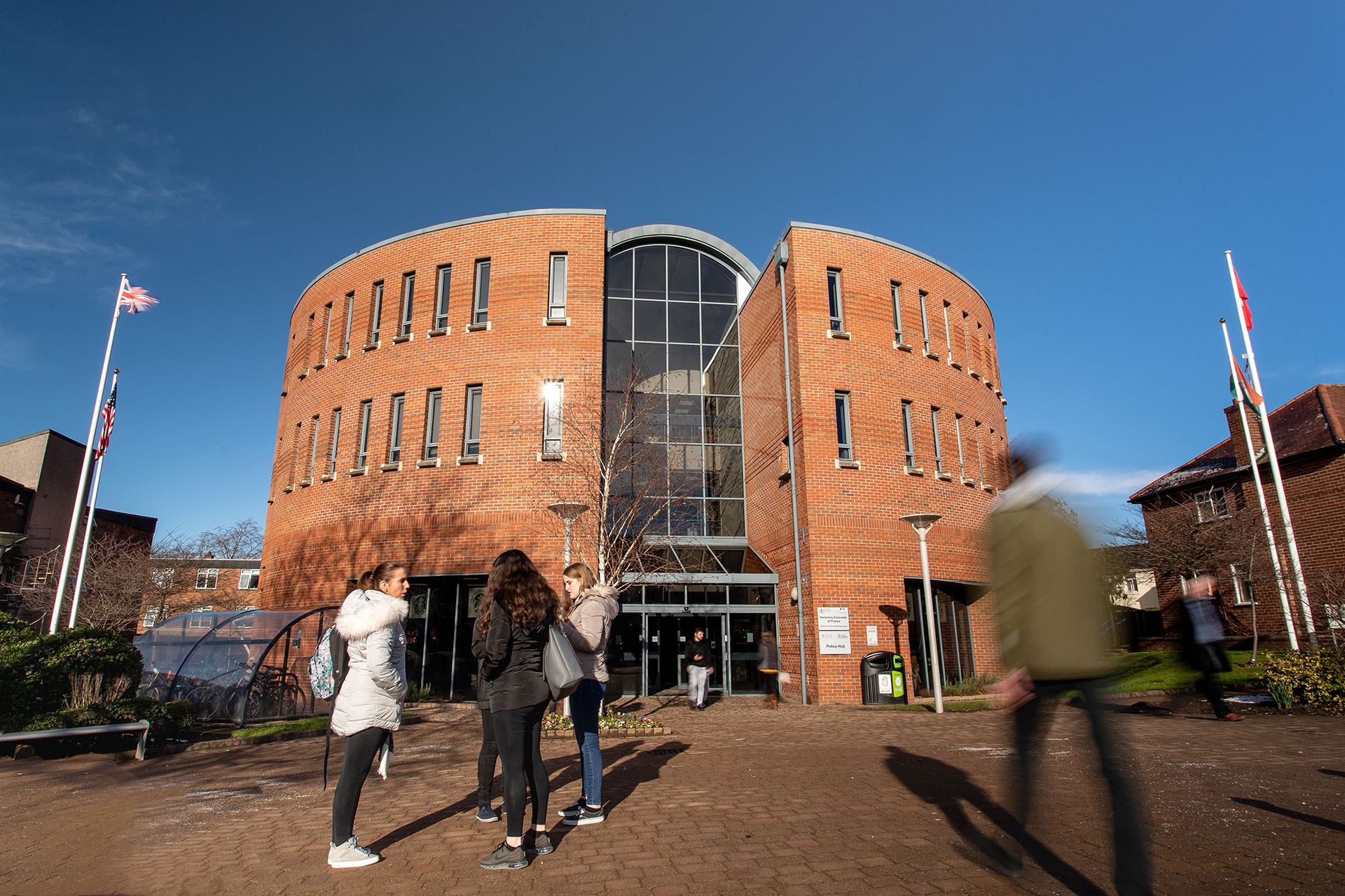 A group of people stand and walk near a modern, curved brick building with large windows at Chester University under a clear blue sky.