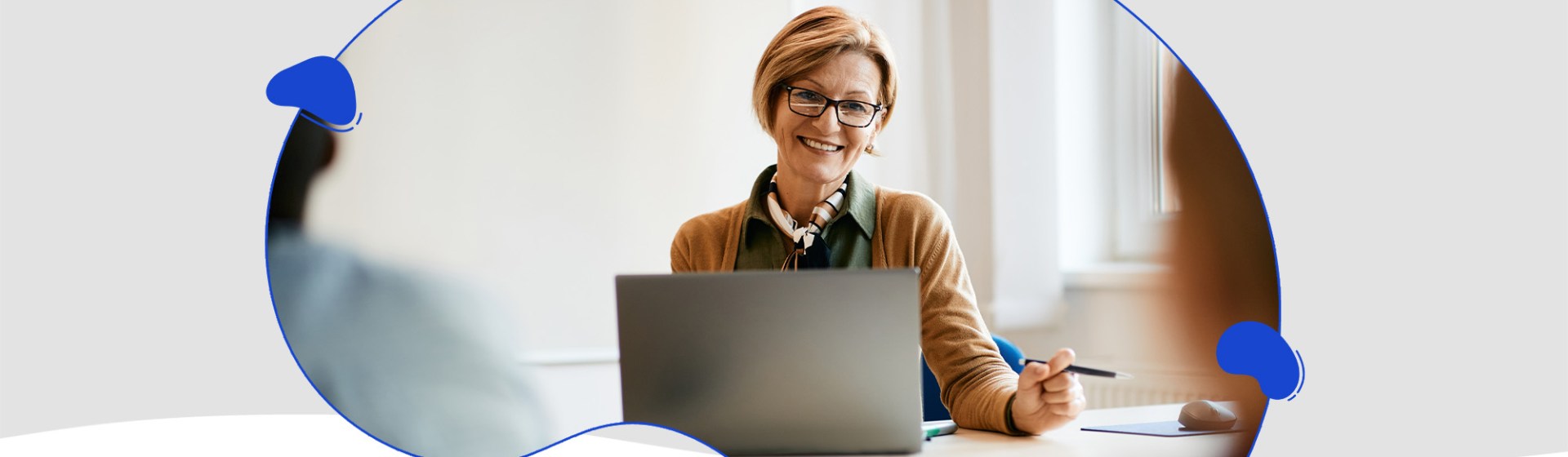 A person with short hair, wearing glasses and a scarf, smiles while working on a laptop at a desk in a bright room, researching ways to upskill your staff for effective AI integration in the classroom.