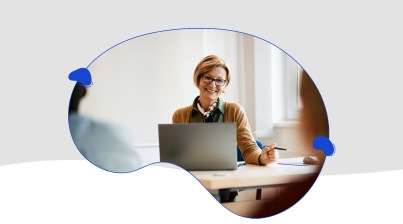 A person with short hair, wearing glasses and a scarf, smiles while working on a laptop at a desk in a bright room, researching ways to upskill your staff for effective AI integration in the classroom.