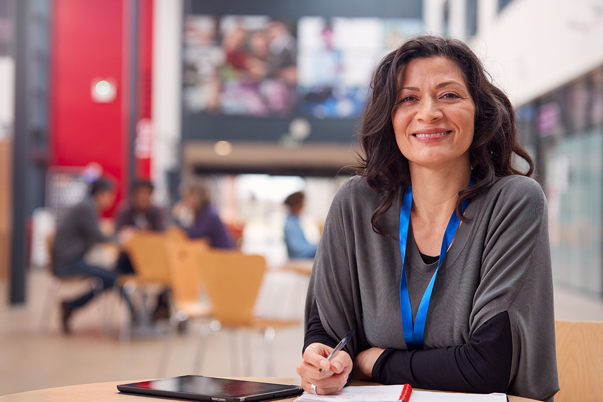 Woman with dark hair and a blue lanyard sits at a table, smiling, with a notebook and tablet in front of her—perhaps reviewing the TeacherMatic Higher Education Pilot Report in a modern indoor setting.