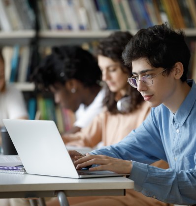Students sitting at a library table, focused on laptops and notebooks, with bookshelves in the background.