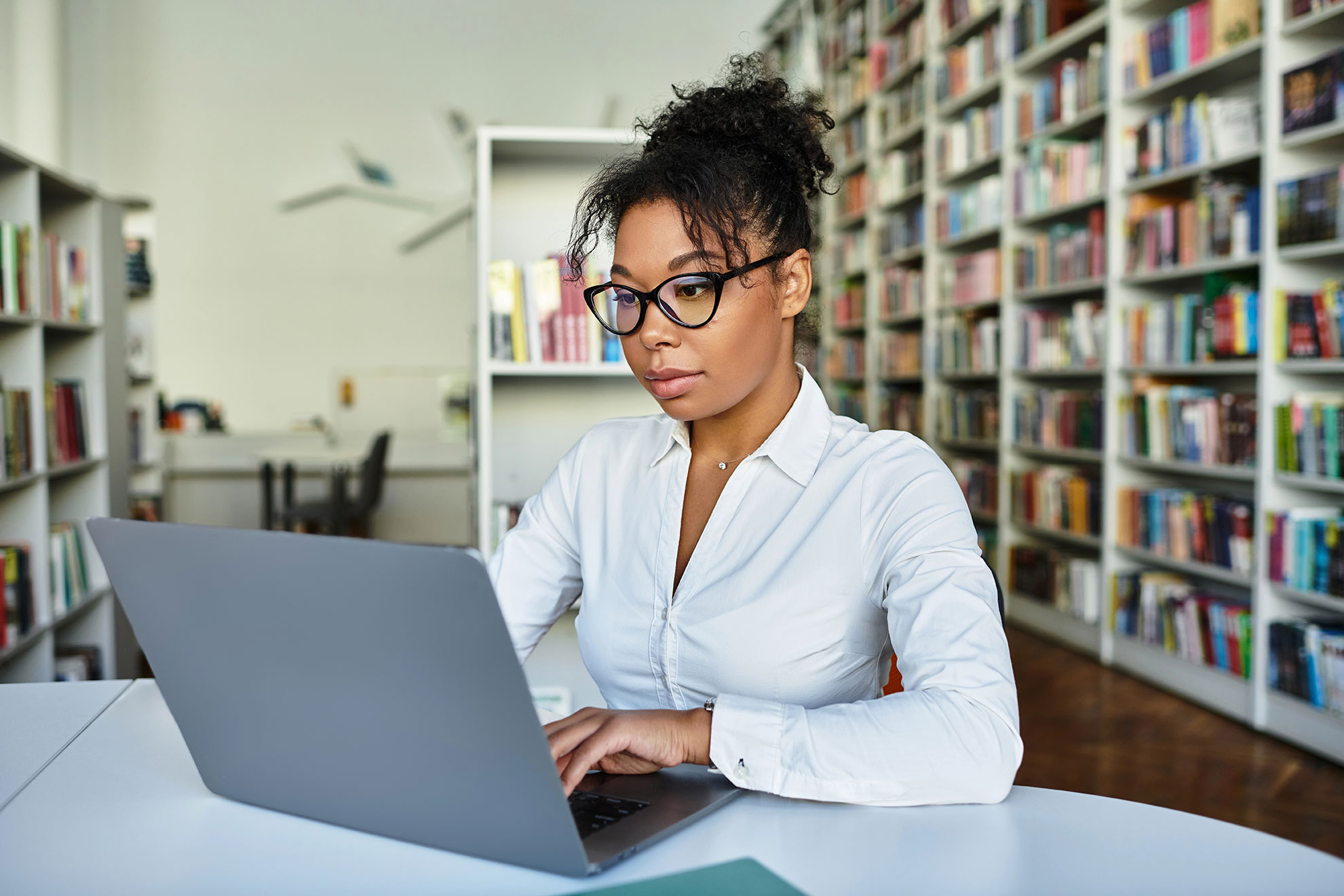 A woman wearing glasses and a white shirt works on a laptop at a table in a library filled with bookshelves, exploring Generative AI tools for educators to enhance responsible AI adoption in universities.