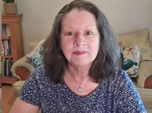 A woman with shoulder-length dark hair and a floral top sits in a room with a couch and bookshelf in the background.