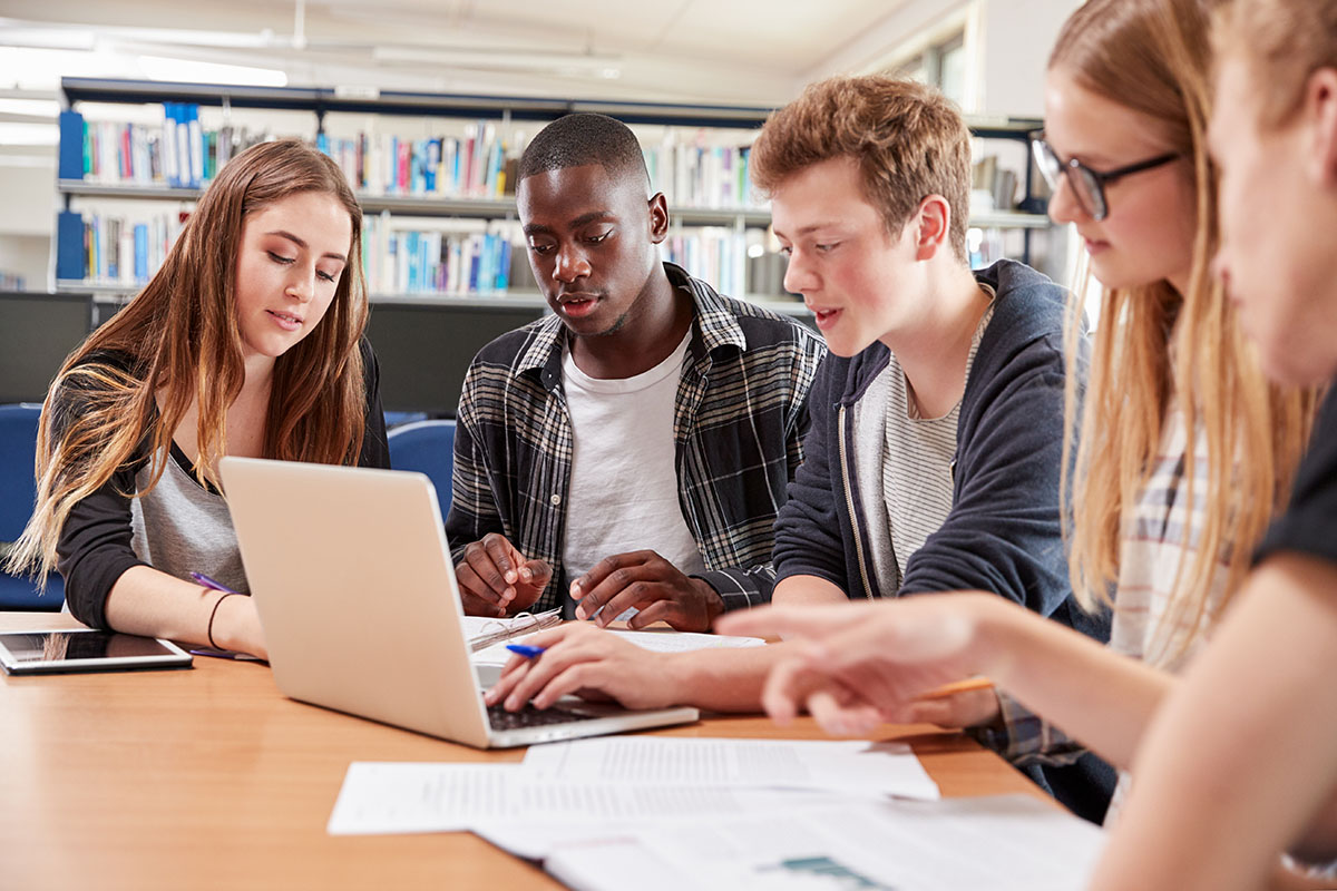 Five students sit around a table in a library, looking at a laptop and documents, collaborating on a group project using insights from the TeacherMatic Higher Education Pilot Report.