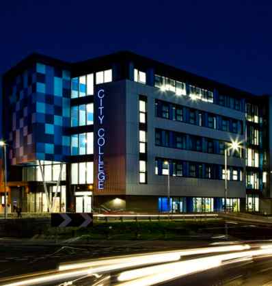 The City College building, a shining example of modern architecture, is illuminated at night with blue and white exterior panels. Light trails from passing cars create a dynamic foreground, making it a perfect case study in urban design and lighting.