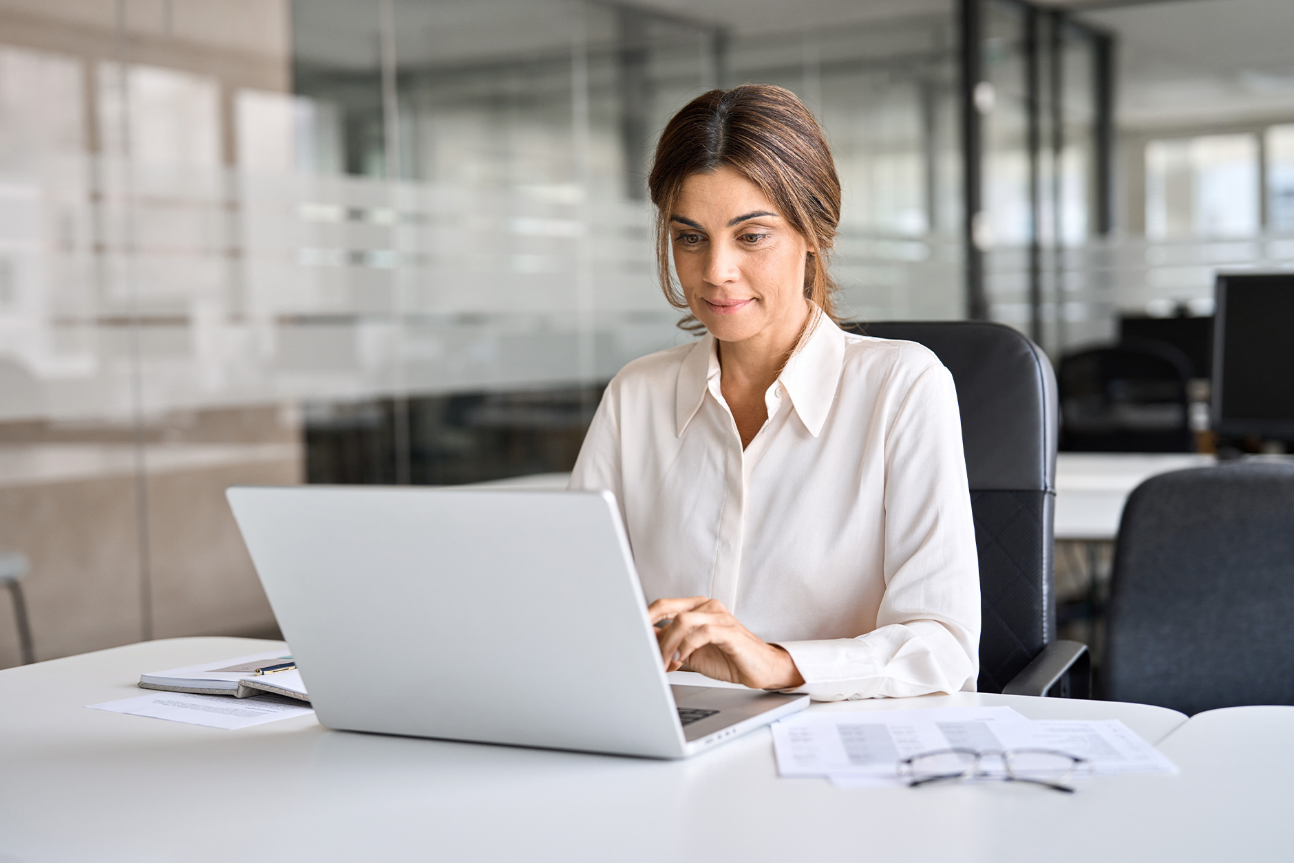 A woman in a white shirt works on a laptop at an office desk with papers nearby.
