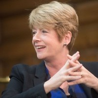 A person with short blonde hair is smiling, facing right. They are wearing a dark blazer with a blue top and have their hands clasped.