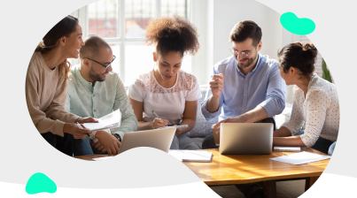 Five people are sitting around a table, engaged in discussion, with laptops and notebooks in front of them.