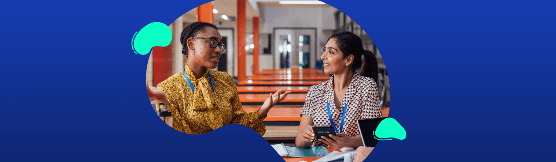Two women sit at a table indoors, talking and smiling with notebooks and pens in front of them against a blue background, capturing a moment from a week in the life of a School Leader.