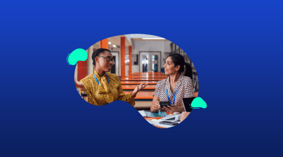 Two women sit at a table indoors, talking and smiling with notebooks and pens in front of them against a blue background, capturing a moment from a week in the life of a School Leader.