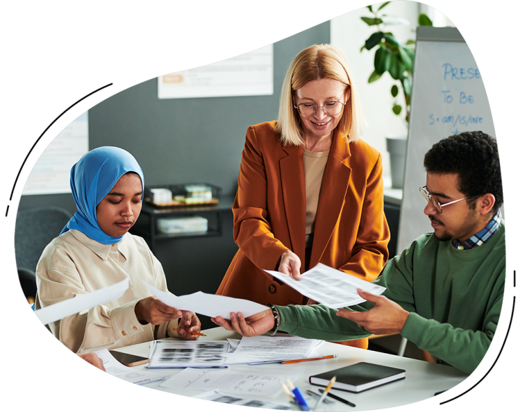 Three people are sitting at a table reviewing and exchanging documents in an office setting, with TeacherMatic Language Teaching Edition materials, papers, and charts spread out in front of them.