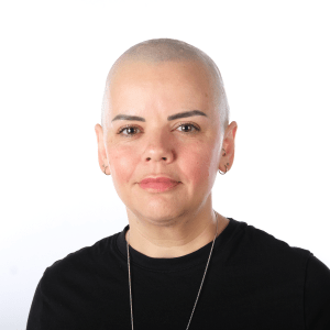 Studio portrait of a bald woman with a neutral expression, wearing a black shirt and a silver necklace against a white background.