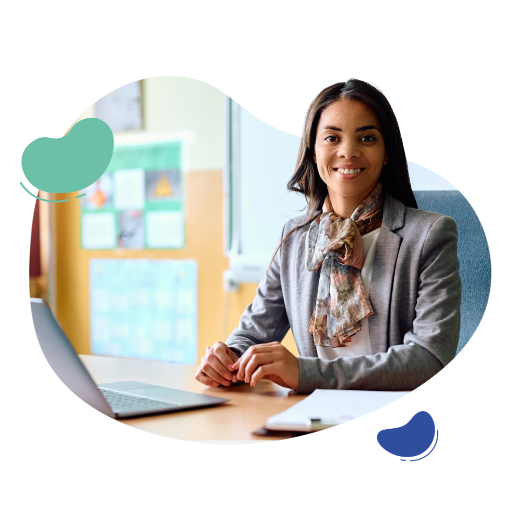 A person in a gray blazer and scarf sits at a desk with a laptop, smiling. There's a blurred background of a classroom with colorful posters.