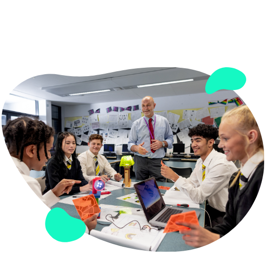 A group of students and a teacher are engaged in a classroom activity around a table with electronic components and laptops. The students are in school uniforms and appear to be discussing their project.