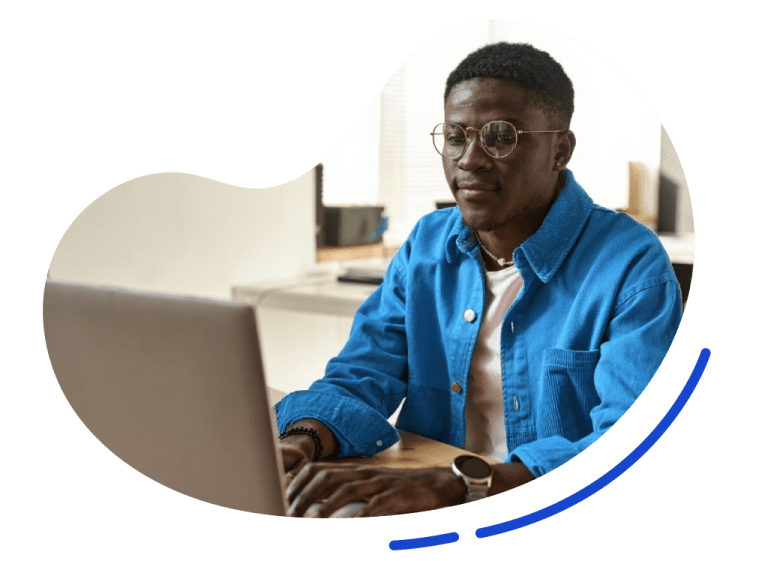 A man wearing glasses and a blue jacket works on a laptop at a desk in a bright room.
