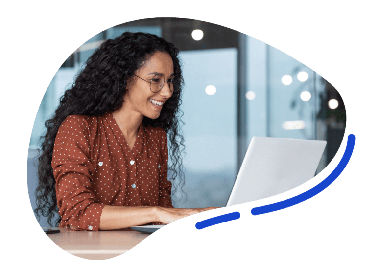 A woman with curly hair and glasses is smiling while working on a laptop at a desk in a modern office setting.