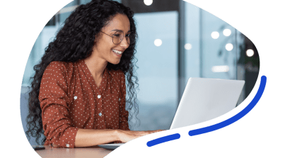 A woman with curly hair and glasses is smiling while working on a laptop at a desk in a modern office setting.