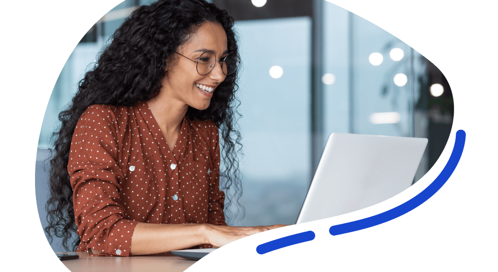 A woman with curly hair and glasses is smiling while working on a laptop at a desk in a modern office setting.